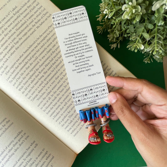 Miniature Tribal legs  made of clay wearing  tribal toe rings and kara an dred kohlapuri chappal in blue skirt with maroon flowers as a  bookmark with Aprajita toor's poem on it.
